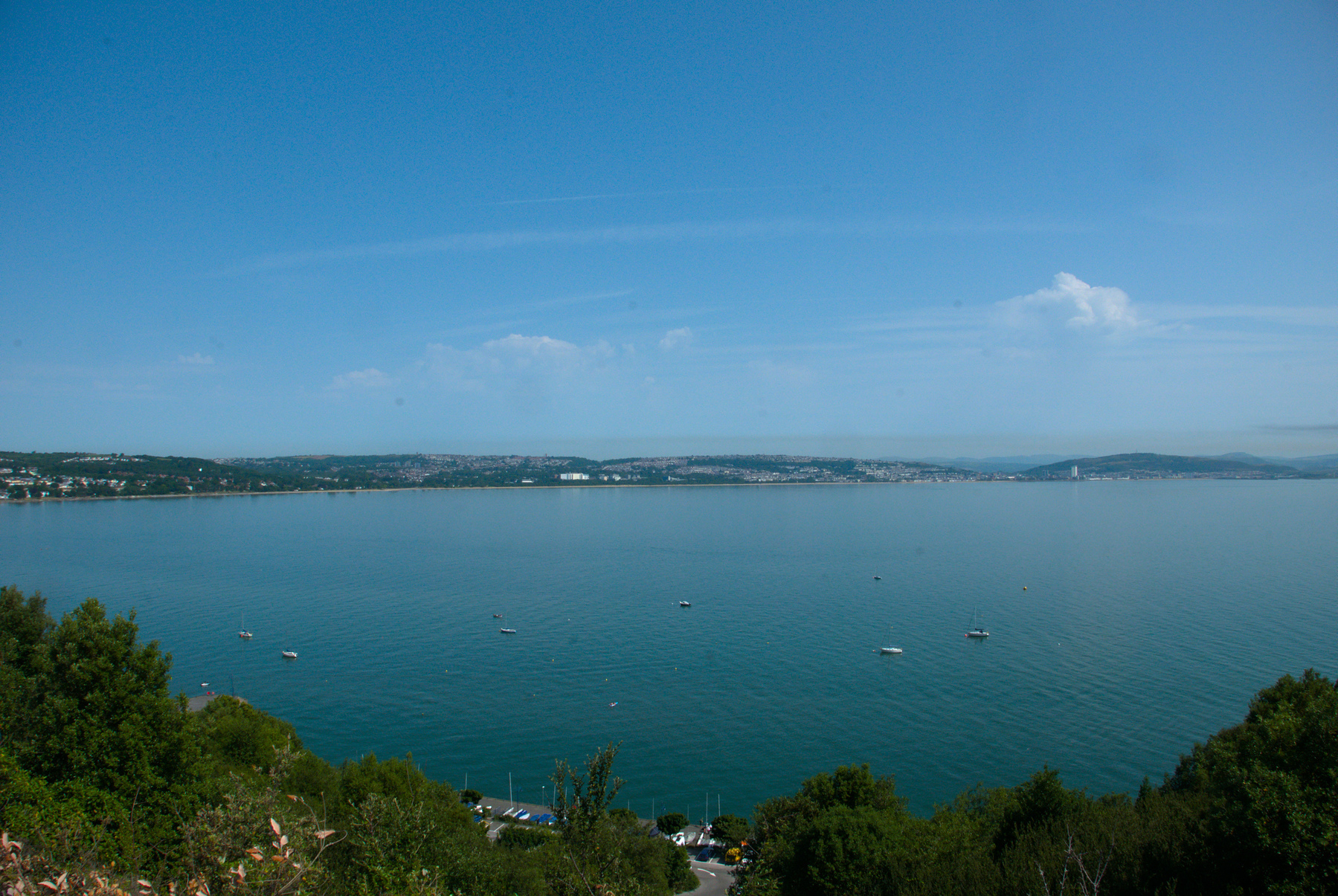 Photograph of a bay with a few scattered small boats and buoys. The sky and water are blue, and the land is green.