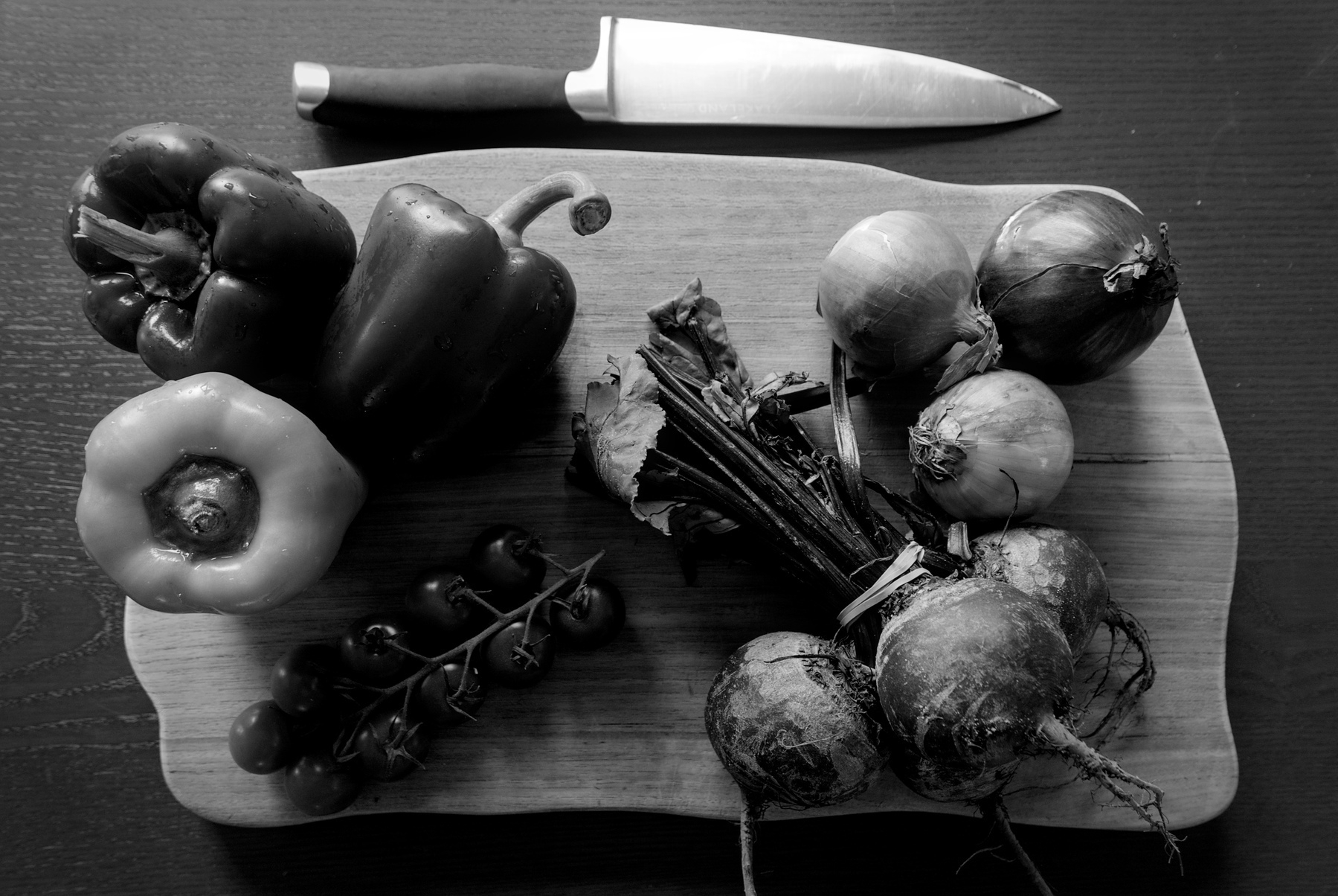 Black-and-white still-life photograph of three bell peppers, three onions, a bunch of beets, and a vine of tomatoes on a cutting board with a chef's knife lying close-by