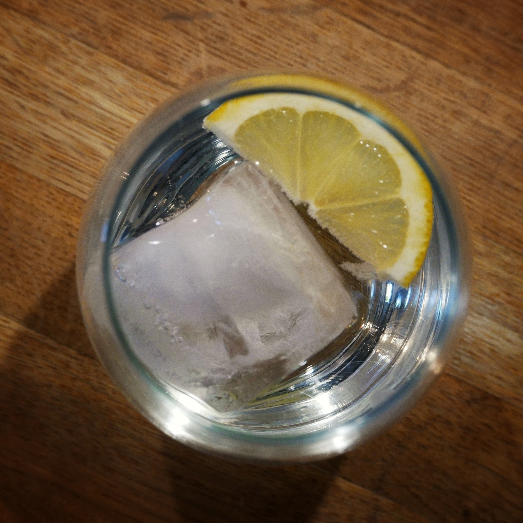 A top-down view of a glass of clear liquid with a large ice cube and wedge of lemon, placed on a wooden surface