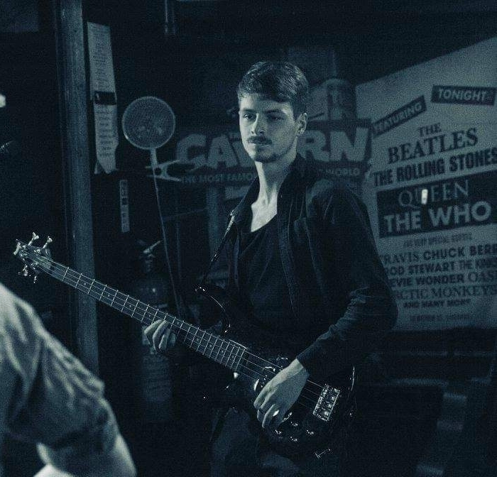 A man playing an electric bass guitar, with a 'Cavern club' poster visible in the background