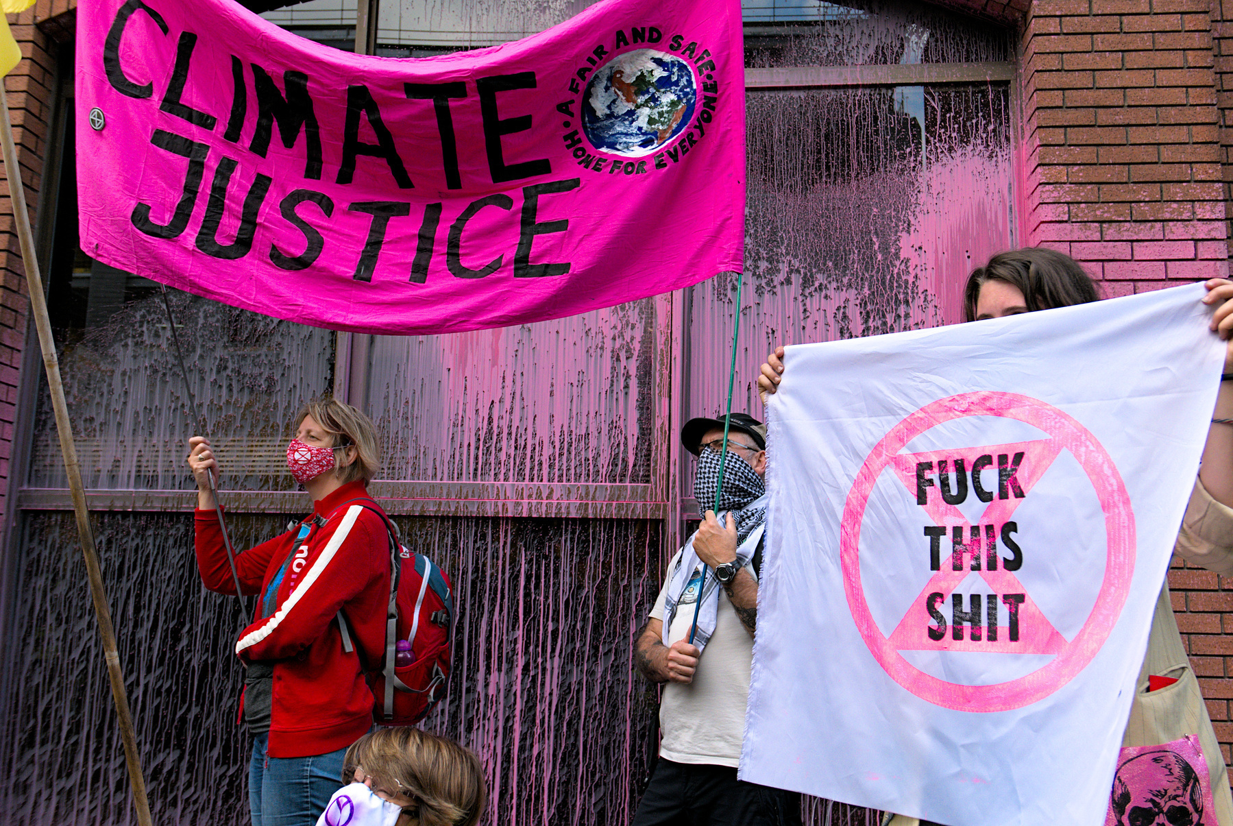 Photograph from a climate protest. Two people wearing covid masks wave a pink banner saying 'climate justice'. Another person holds a banner with a pink XR logo and the words 'fuck this shit'. There is pink painted splashed across the walls of the building behind