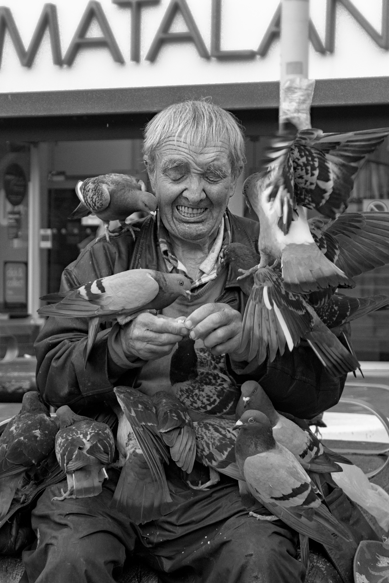 Black and white photo, portrait view of a man sitting outside a shop feeding pigeons, with many pigeons sitting and climbing all over him