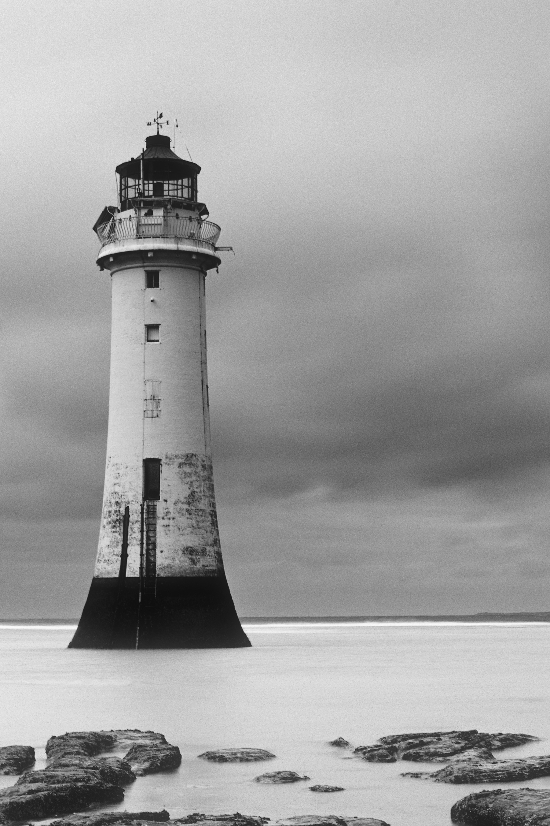 A black and white photo of a lighthouse with some rocks in the foreground. The clouds are slightly dark but the sea looks still.